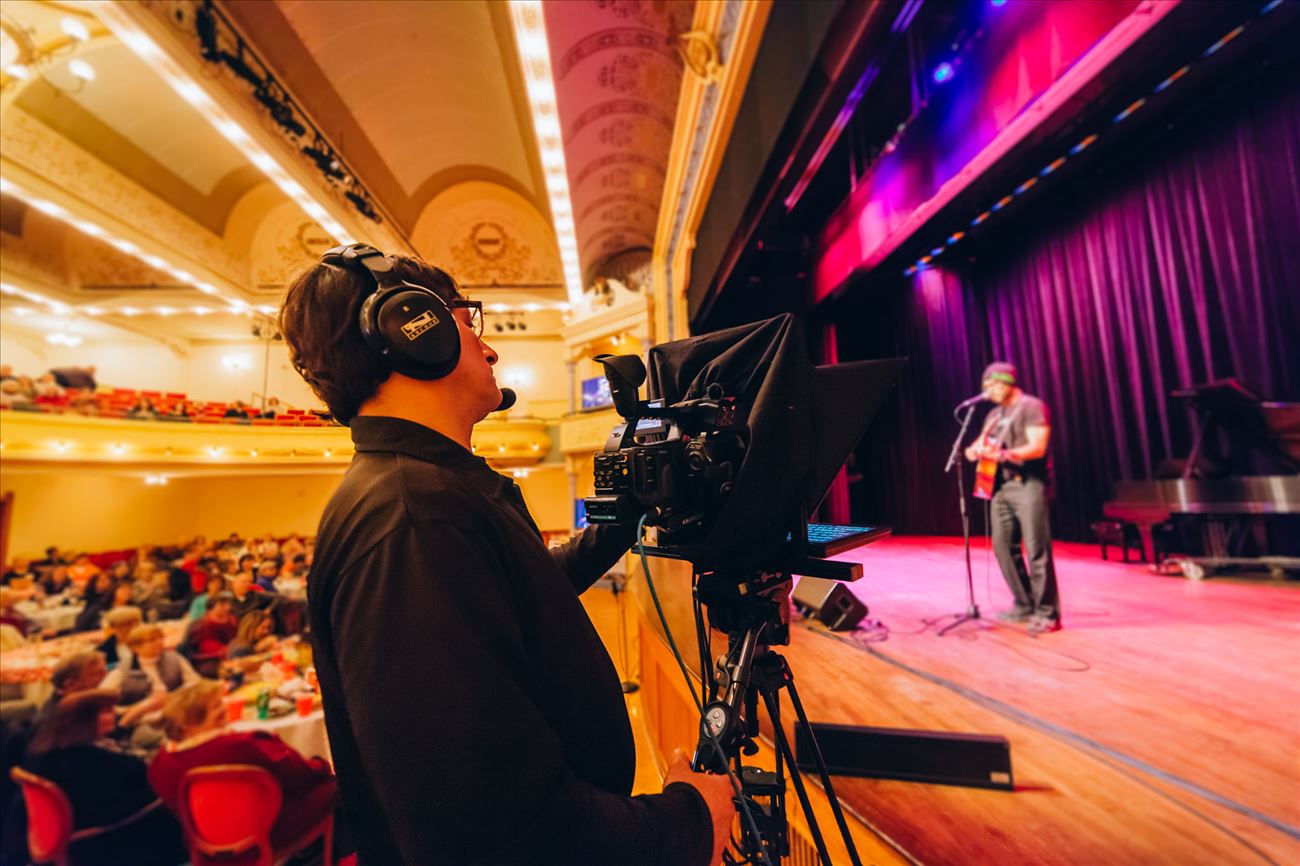 A camera operator films a guitar player while an audience looks on